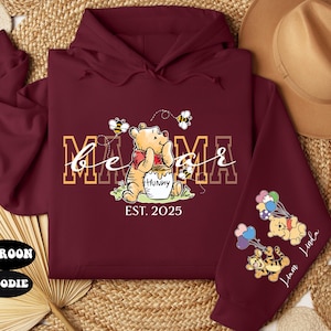 May include: Maroon hoodie with a Winnie the Pooh design. The text "be a mama est. 2025" is printed on the front. The sleeves feature Winnie the Pooh characters with balloons.