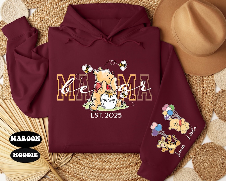 May include: Maroon hoodie with a Winnie the Pooh design. The text "be a mama est. 2025" is printed on the front. The sleeves feature Winnie the Pooh characters with balloons.