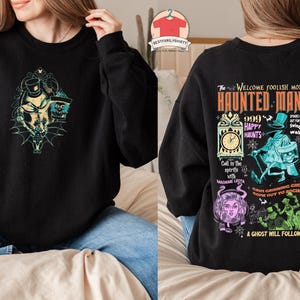 May include: Black sweatshirt with a colorful graphic print of the Haunted Mansion attraction at Disneyland. The print features the text "The Welcome Foolish Mortals Haunted Mansion 999 Happy Haunts" and various characters from the attraction.