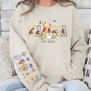 May include: A beige sweatshirt with the text "be a mama est. 2025" and a cartoon Winnie the Pooh holding a honey pot. The sleeves have a line of cartoon Winnie the Pooh characters with balloons.