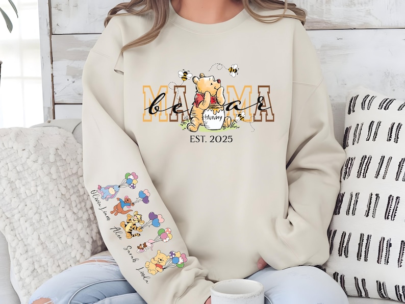 May include: A beige sweatshirt with the text "be a mama est. 2025" and a cartoon Winnie the Pooh holding a honey pot. The sleeves have a line of cartoon Winnie the Pooh characters with balloons.