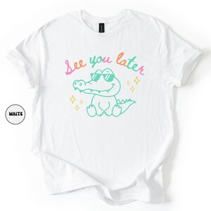 Puede incluir: Camiseta blanca con un cocodrilo de dibujos animados con gafas de sol y el texto "See you later" en rosa y verde. El cocodrilo es verde con detalles amarillos. Prenda de vestir informal.