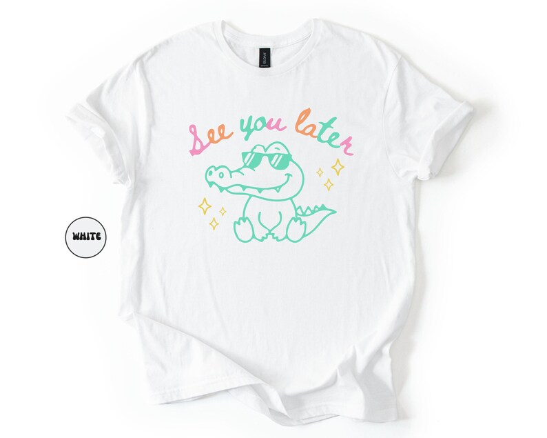 Puede incluir: Camiseta blanca con un cocodrilo de dibujos animados con gafas de sol y el texto "See you later" en rosa y verde. El cocodrilo es verde con detalles amarillos. Prenda de vestir informal.