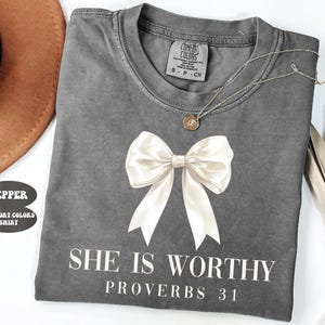 May include: A gray Comfort Colors t-shirt with a large white bow graphic and the text "SHE IS WORTHY PROVERBS 31". A silver necklace with a small pendant is draped across the shirt.