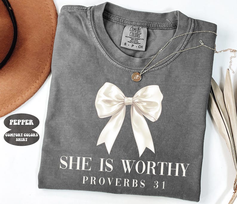 May include: A gray Comfort Colors t-shirt with a large white bow graphic and the text "SHE IS WORTHY PROVERBS 31". A silver necklace with a small pendant is draped across the shirt.