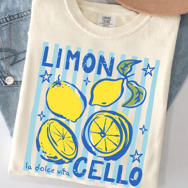 May include: Cream-colored t-shirt with a lemon-themed graphic. The design features the words "LIMONCELLO" in blue, along with illustrations of lemons and the phrase "la dolce vita". The shirt has a crew neck and short sleeves.