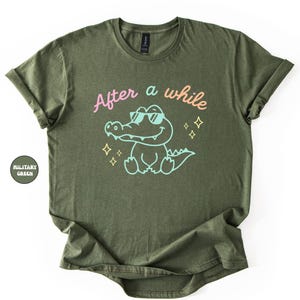 Puede incluir: Camiseta verde oliva con el texto "After a while" sobre un cocodrilo de dibujos animados con gafas de sol. El cocodrilo est&aacute; delineado en azul claro, con detalles en rosa y amarillo. La camiseta est&aacute; doblada.