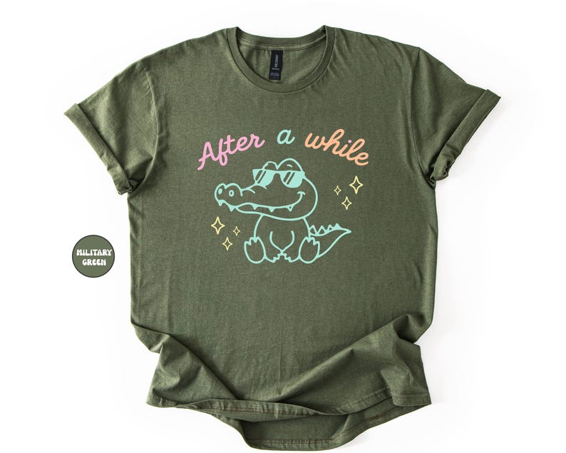 Puede incluir: Camiseta verde oliva con el texto "After a while" sobre un cocodrilo de dibujos animados con gafas de sol. El cocodrilo est&aacute; delineado en azul claro, con detalles en rosa y amarillo. La camiseta est&aacute; doblada.