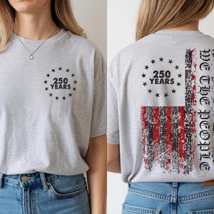 Może przedstawiać: Jasnoszara koszulka z napisem "250 YEARS" w okręgu z gwiazd na przodzie i z tyłu. Tył przedstawia postarzany wz&oacute;r amerykańskiej flagi ze słowami "We The People".