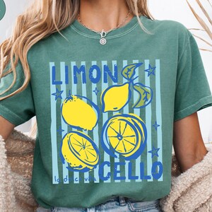 May include: A light green t-shirt with a lemon-themed graphic. The design features yellow lemons, blue stripes, and the words "LIMONCELLO" in blue. The shirt has a relaxed fit and short sleeves.