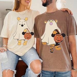 May include: A cream-colored t-shirt featuring a cartoon bear dressed as a ghost holding a pumpkin bucket, and a brown t-shirt featuring a cartoon tiger dressed as a ghost holding a pumpkin bucket, both with Mickey Mouse ear silhouettes.