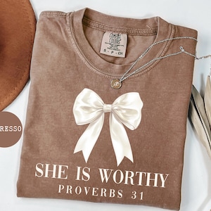 May include: Espresso-colored t-shirt with a large white bow graphic and the text "SHE IS WORTHY PROVERBS 31". The shirt has a crew neck and a small tag. A silver necklace with a pendant is draped across the shirt.