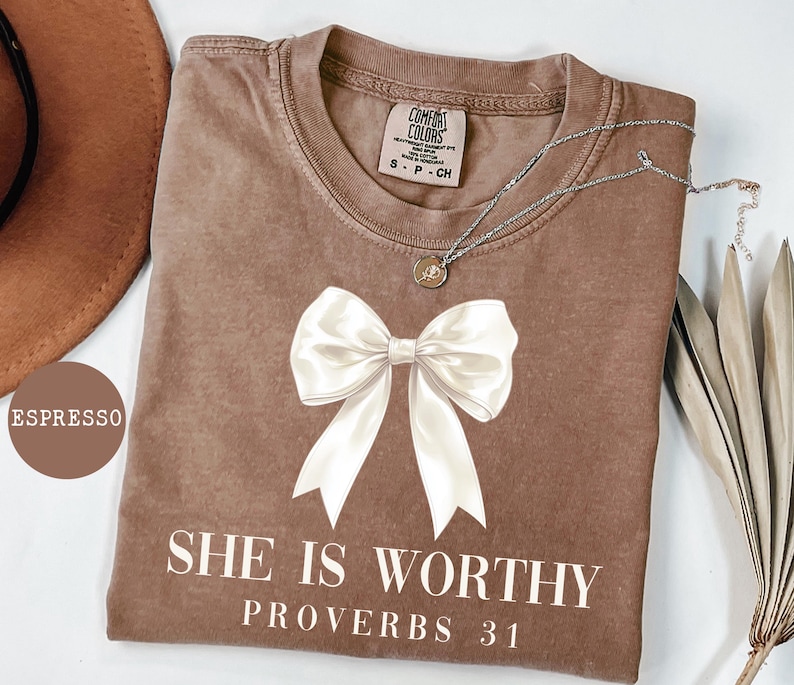 May include: Espresso-colored t-shirt with a large white bow graphic and the text "SHE IS WORTHY PROVERBS 31". The shirt has a crew neck and a small tag. A silver necklace with a pendant is draped across the shirt.