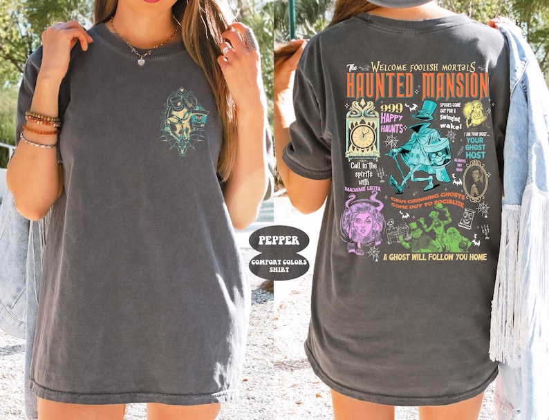 May include: A gray t-shirt with a graphic print of the Haunted Mansion attraction at Disneyland. The graphic includes the text "The Welcome Foolish Mortals Haunted Mansion", "999 Happy Haunts", "Your Ghost Host", and "A Ghost Will Follow You Home".