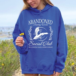 May include: Royal blue hoodie with the text "ABANDONED Hunting Wives Social Club" and a graphic of a flying duck. The text "HELLO HUNTING SEASON, GOODBYE HUSBAND" is also printed on the hoodie.