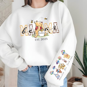 May include: White sweatshirt with a Winnie the Pooh inspired design. The text "be a mama" is printed in brown and yellow letters. The text "EST. 2025" is printed below the design. The sleeve has a design of Winnie the Pooh characters with their names printed below.