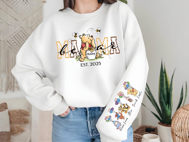 May include: White sweatshirt with a Winnie the Pooh inspired design. The text "be a mama" is printed in brown and yellow letters. The text "EST. 2025" is printed below the design. The sleeve has a design of Winnie the Pooh characters with their names printed below.