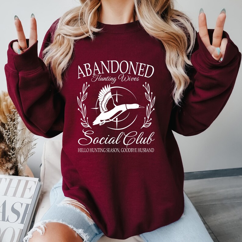 May include: A maroon sweatshirt with white text and a graphic of a flying duck. The text reads "ABANDONED Hunting Wives Social Club" with the phrase "Hello Hunting Season, Goodbye Husband" below the graphic.