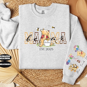 May include: Gray sweatshirt with a Winnie the Pooh design. The text "Mama Bear Est. 2025" is printed on the front. The sleeve has a design of Winnie the Pooh characters with balloons and the text "Alex Sarah Luna".