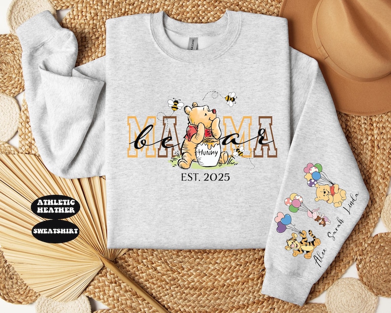 May include: Gray sweatshirt with a Winnie the Pooh design. The text "Mama Bear Est. 2025" is printed on the front. The sleeve has a design of Winnie the Pooh characters with balloons and the text "Alex Sarah Luna".