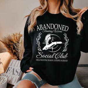 May include: Black sweatshirt with white text and a graphic of a flying duck. The text reads "ABANDONED Hunting Wives Social Club" with the phrase "Hello Hunting Season, Goodbye Husband" below the graphic.