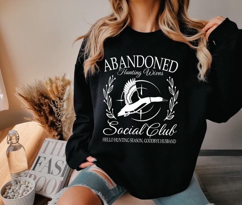 May include: Black sweatshirt with white text and a graphic of a flying duck. The text reads "ABANDONED Hunting Wives Social Club" with the phrase "Hello Hunting Season, Goodbye Husband" below the graphic.