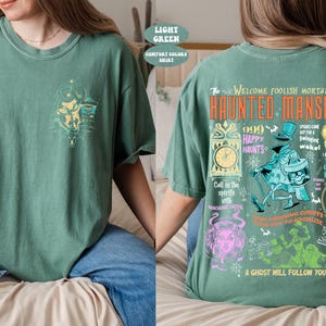 May include: A light green t-shirt with a graphic print of the Haunted Mansion attraction from Disneyland. The text on the shirt reads "The Welcome Foolish Mortals Haunted Mansion 999 Happy Haunts".