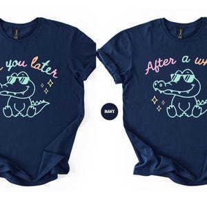 Puede incluir: Camisetas azul marino con un cocodrilo de dibujos animados con gafas de sol. Una camiseta dice "See you later" y la otra "After a while" en escritura colorida. Ambas camisetas tienen un c&iacute;rculo azul marino con la palabra "NAVY".