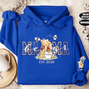 May include: Blue hooded sweatshirt with a Winnie the Pooh character holding a pot of honey. The text "be a mama" is printed in gold with bees and the year "EST. 2025" below.