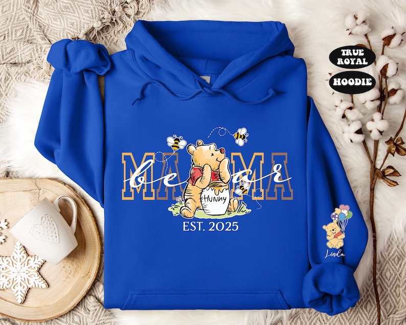 May include: Blue hooded sweatshirt with a Winnie the Pooh character holding a pot of honey. The text "be a mama" is printed in gold with bees and the year "EST. 2025" below.