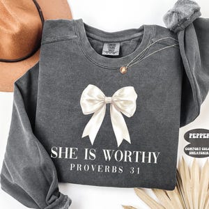 May include: A dark gray sweatshirt with a large white bow graphic and the text "SHE IS WORTHY PROVERBS 31" in white. The sweatshirt has long sleeves and a crew neck. A gold necklace is visible.