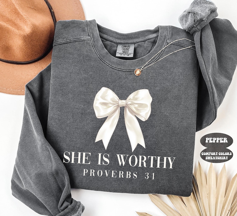 May include: A dark gray sweatshirt with a large white bow graphic and the text "SHE IS WORTHY PROVERBS 31" in white. The sweatshirt has long sleeves and a crew neck. A gold necklace is visible.