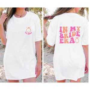 May include: White t-shirt with a pink and orange graphic that says "In My Bride Era" with a diamond ring graphic. The front of the shirt has a small diamond ring graphic with the text "Mrs. Johnson".