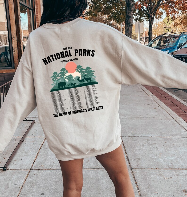 May include: A beige sweatshirt with a graphic print on the back that says "Keep Our National Parks Pristine & Unspoiled" with a green forest scene and the text "The Heart of America's Wildlands".