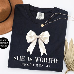 May include: Black Comfort Colors crew neck t-shirt with a large white bow graphic and the text "SHE IS WORTHY PROVERBS 31". A gold necklace and a brown hat are also visible.