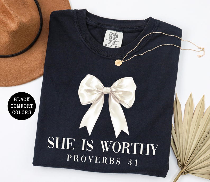 May include: Black Comfort Colors crew neck t-shirt with a large white bow graphic and the text "SHE IS WORTHY PROVERBS 31". A gold necklace and a brown hat are also visible.