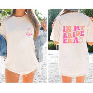 May include: White t-shirt with a pink and orange graphic that says "In My Bride Era" with a diamond ring on the back. The front of the shirt has a pink and orange ring with the text "Mrs. Johnson" on a white t-shirt.