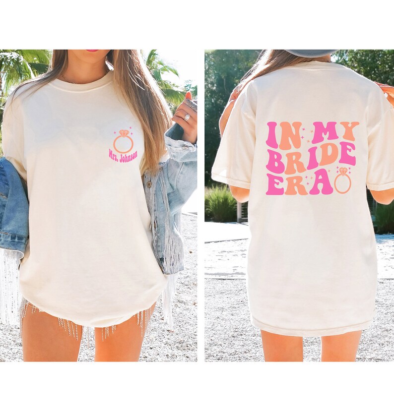 May include: White t-shirt with a pink and orange graphic that says "In My Bride Era" with a diamond ring on the back. The front of the shirt has a pink and orange ring with the text "Mrs. Johnson" on a white t-shirt.