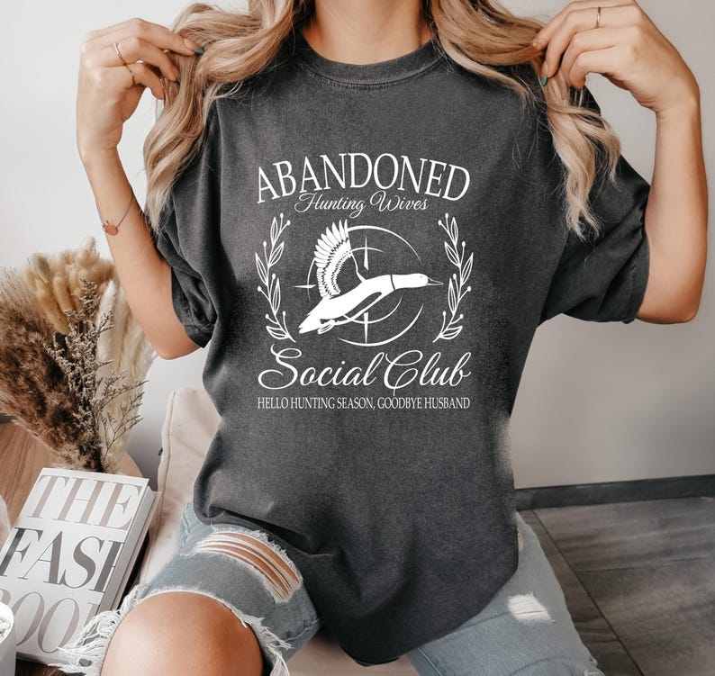 May include: Dark gray t-shirt with the text "ABANDONED Hunting Wives Social Club" in white script. A white graphic of a flying duck is centered within a circle. The text "HELLO HUNTING SEASON, GOODBYE HUSBAND" is below.