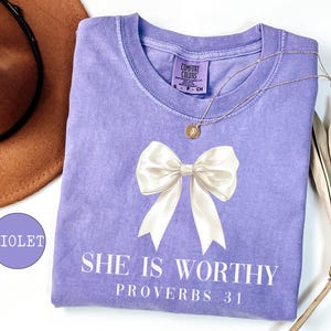 May include: A violet Comfort Colors t-shirt with a large white bow graphic and the text "SHE IS WORTHY PROVERBS 31". A gold necklace and a brown hat are also visible.