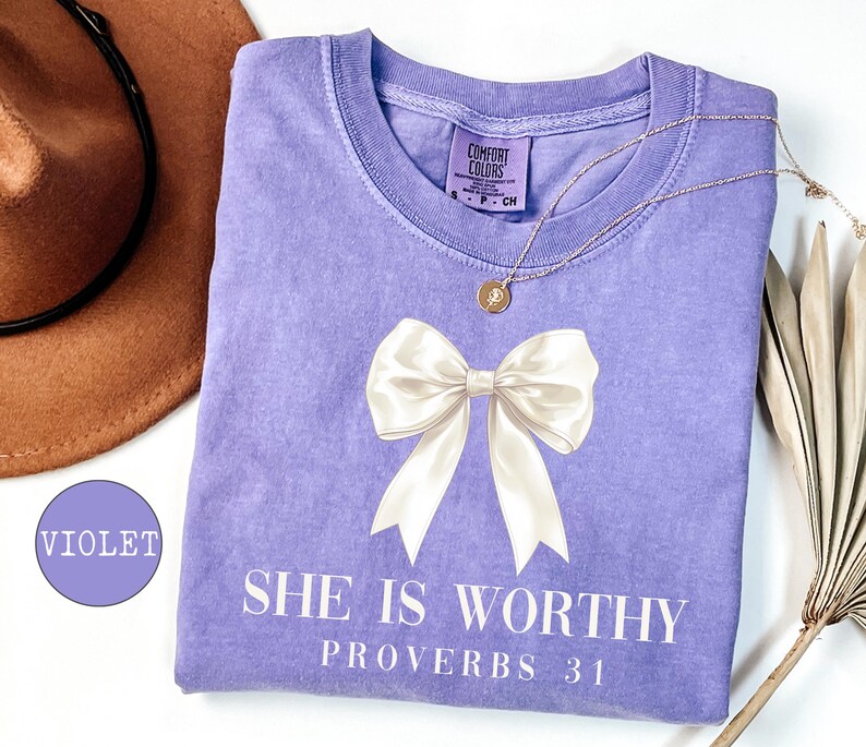 May include: A violet Comfort Colors t-shirt with a large white bow graphic and the text "SHE IS WORTHY PROVERBS 31". A gold necklace and a brown hat are also visible.