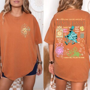 May include: A burnt orange t-shirt with a graphic print of a haunted mansion. The text on the shirt reads "The Welcome Foolish Mortals Haunted Mansion 666 Happy Haunts Call in the spirits with a midnight light A ghost will follow you home."