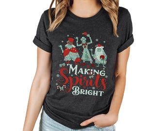Puede incluir: Una camiseta gris oscuro de manga corta con un gráfico de tres personajes verdes decorados con luces navideñas y gorros. El texto "MAKING SPIRITS BRIGHT" está impreso en rojo y blanco debajo de los personajes, rodeado de copos de nieve.