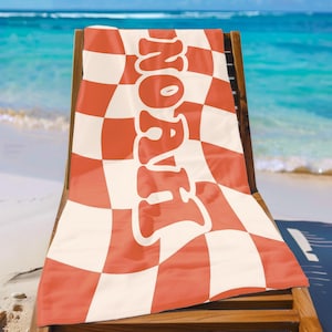 May include: A beach towel with a red and white checkered pattern and the name "NOAH" in large, white, stylized letters. The towel is draped over a wooden beach chair, with the ocean and sky in the background.