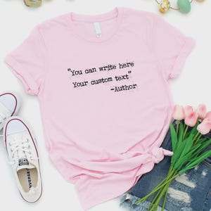 Może przedstawiać: Jasnor&oacute;żowa koszulka z czarnym napisem "You can write here Your custom text". Koszulka jest zawiązana na dole i połączona z dżinsowymi szortami, białymi trampkami i r&oacute;żowymi tulipanami.