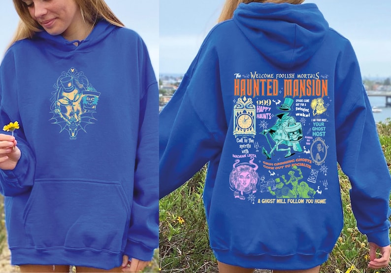 May include: A royal blue hoodie with a graphic print of the Haunted Mansion attraction at Disneyland. The print features a clock, ghosts, and the text "The Welcome Foolish Mortals Haunted Mansion 1969 Happy Haunts".