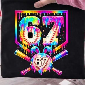Puede incluir: Bolso tote negro con un diseño colorido de béisbol. El diseño incluye el número 67 en forma de béisbol, con efectos de pintura de arcoíris goteando. Bate de béisbol cruzados y una pelota de béisbol con el número 67 completan el diseño.