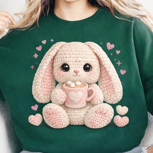May include: A dark green sweatshirt featuring a cute, crocheted bunny holding a pink mug of hot chocolate. The bunny has pink inner ears and paws. Pink hearts and cross accents are scattered around the bunny.