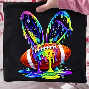 May include: Black tote bag featuring a colorful graphic of a football with bunny ears. The football is orange with white stripes and laces. The bunny ears and dripping paint are a rainbow of colors.