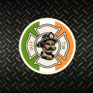 以下が含まれることがあります： 緑、白、オレンジのカラーリングの円形のステッカー。ステッカーには、口ひげとヘルメットをつけた消防士、IAFFという文字と718という数字の入った十字架が描かれている。ステッカーは黒い背景に貼られている。